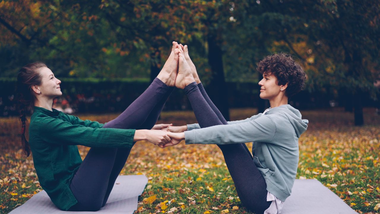 services-02 Two women practicing partner yoga outdoors on a fall day, surrounded by colorful leaves.